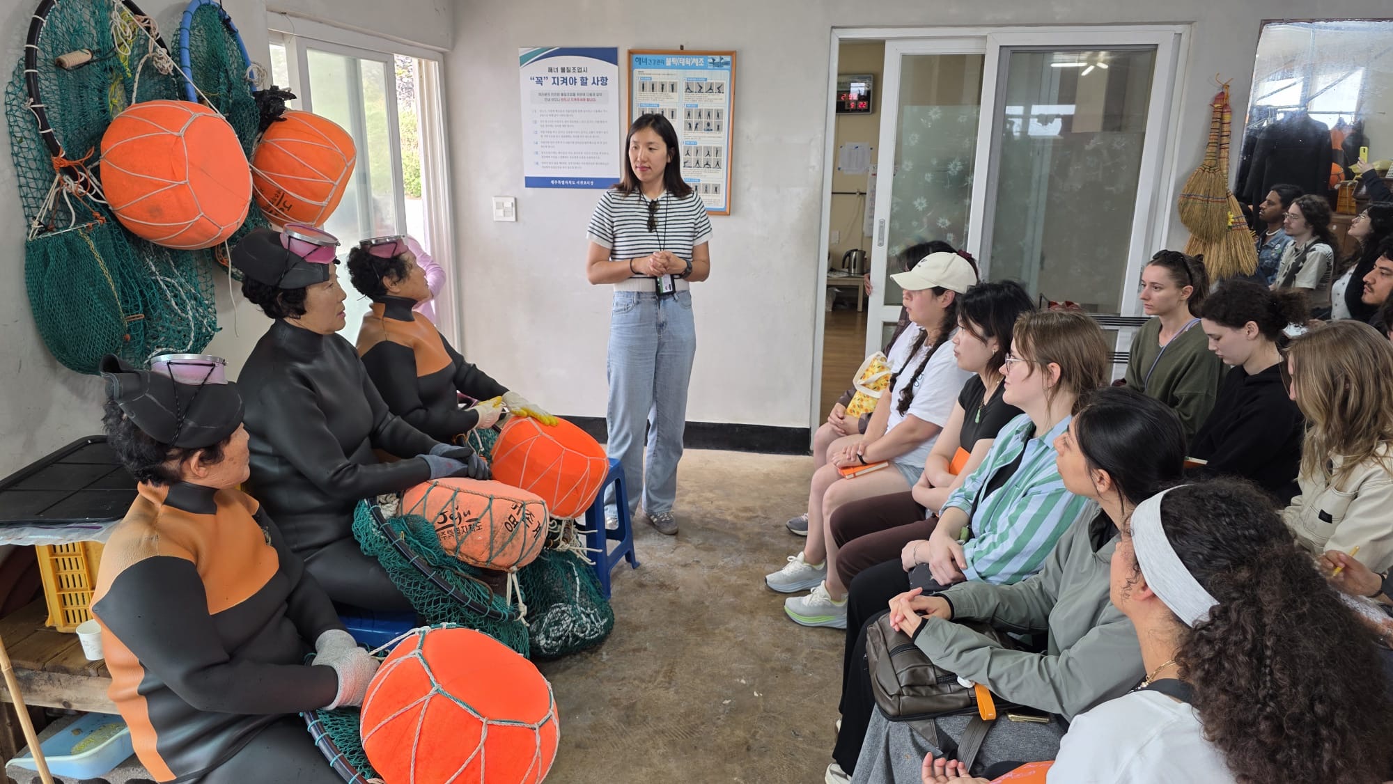 A guide introduces the haenyeo to guests, the sea women seated with their tewak floats